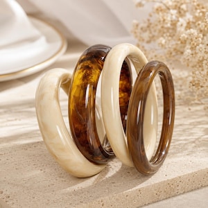 May include: A set of four circular bangle bracelets featuring a smooth, polished finish. Two bracelets are a solid cream colour with subtle marbling, while the other two display a rich, translucent dark brown tortoiseshell pattern with depth.