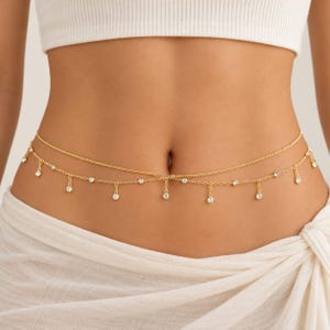 May include: A gold-toned waist chain featuring two delicate layered chains. The lower chain is adorned with evenly spaced, small, clear round crystals that hang as individual charms along the length of the chain, resting against the midsection.