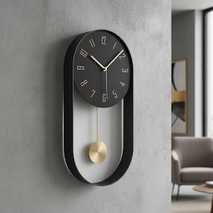 May include: A wall clock with a black oval frame and a circular black face featuring gold-coloured numerals and hands. A thin gold rod extends downwards from the clock face to a circular gold pendulum, suspended within the open lower section.