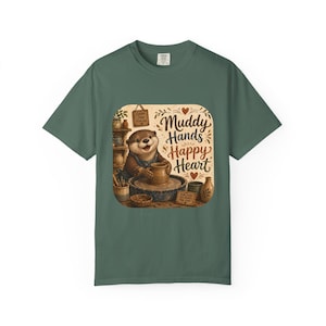 May include: A dark sage green t-shirt featuring a graphic of an otter working at a pottery wheel. The illustration includes pottery tools, bowls, and the text "Muddy Hands Happy Heart" in a decorative brown font, surrounded by small red hearts.