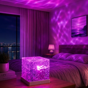 May include: A square, textured glass lamp emitting a vibrant purple light that projects a rippling, water-like pattern onto the surrounding walls and ceiling. The lamp sits on a white base and is positioned on a wooden surface in a bedroom.