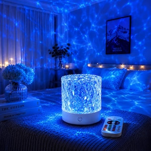 May include: A cylindrical crystal-textured lamp with a white base and a power button, emitting a bright blue light that projects a water-like pattern onto the surrounding surfaces. A small rectangular remote control with multiple buttons sits nearby.