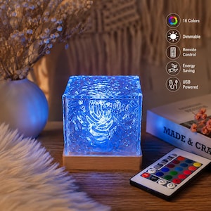 May include: A square, textured glass lamp emitting a blue light, set on a light wood base. Beside it lies a remote control with colorful buttons. Text icons indicate features: 16 Colors, Dimmable, Remote Control, Energy Saving, and USB Powered.