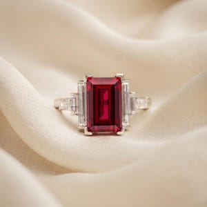 May include: A silver-toned ring featuring a large, rectangular, emerald-cut deep red gemstone at the center. The center stone is flanked on each side by two clear, rectangular baguette-cut stones set in a metallic band with a polished finish.