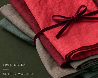May include: A stack of four folded linen napkins in red, burgundy, taupe, and dark green, tied together with a thin dark red velvet ribbon. The text on the image reads 100% LINEN, SOFTLY WASHED, MUTED TONES.