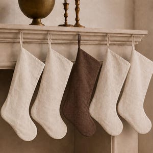 May include: Five hanging Christmas stockings suspended from a mantelpiece. Four stockings are a light beige colour, while the centre stocking is a dark brown. Each stocking features a simple loop at the top for hanging and a textured fabric finish.