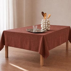 May include: A rectangular table covered with a solid, dark terracotta-colored tablecloth. On the table sits a silver-toned metal tray, a clear glass tumbler, and a white, textured, bulbous ceramic utensil holder containing three wooden spoons.