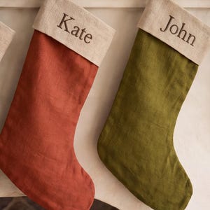 Könnte beinhalten: Zwei Weihnachtssocken hängen nebeneinander. Die linke Socke ist rostorange und hat einen beigen Umschlag, der mit dem Namen Kate bestickt ist. Die rechte Socke ist olivgrün und hat einen beigen Umschlag, der mit dem Namen John bestickt ist.
