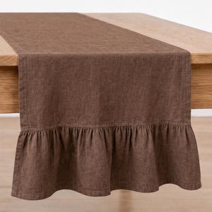 Puede incluir: Un camino de mesa de color marrón oscuro sólido con una superficie de tela texturizada y un borde con volantes fruncidos en la parte inferior. El camino de mesa está drapeado sobre una superficie de madera clara, mostrando el dobladillo recto.
