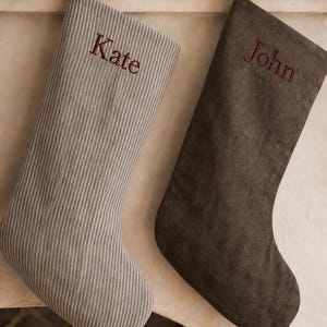 May include: Two holiday stockings hanging from hooks. The left stocking is light grey with thin white vertical stripes and the name Kate embroidered in dark red. The right stocking is solid dark brown with the name John embroidered in dark red.