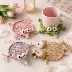 Peut inclure: Quatre dessous de verre ronds en crochet en forme d'animaux, incluant une créature rose, une grenouille verte avec une langue rose, une souris grise aux oreilles rondes et un écureuil beige avec une queue touffue. Un mug rose est posé dessus.