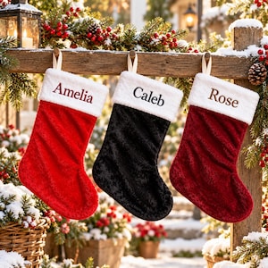 May include: Three plush Christmas stockings hanging from a wooden beam. The left stocking is bright red with the name Amelia embroidered in red. The middle is black with the name Caleb in black. The right is dark red with the name Rose in brown.