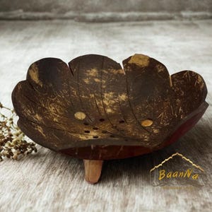 Puede incluir: Un cuenco de cáscara de coco de color marrón oscuro con forma de flor y bordes festoneados. El interior presenta una serie de pequeños orificios de drenaje circulares y una textura fibrosa natural. El cuenco se apoya sobre tres pequeñas patas de madera.