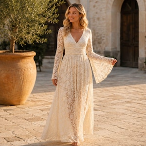 Puede incluir: Un vestido largo de encaje color crema con un escote en V profundo, mangas largas con puños acampanados y una cintura entallada. La prenda está confeccionada con un intrincado patrón de encaje floral que se extiende desde el corpiño al dobladillo.