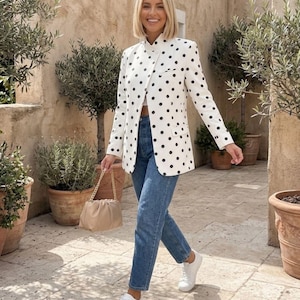 May include: A white blazer with black polka dots, a high collar, and long sleeves, paired with blue denim jeans, white sneakers, and a beige ruched handbag with a gold chain strap. The blazer features a button-front closure and two side pockets.