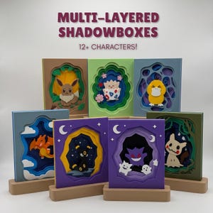 May include: A collection of multi-layered shadowboxes featuring various cartoon characters. Each rectangular frame displays a unique scene with depth, set on a light wooden base. The text at the top reads MULTI-LAYERED SHADOWBOXES 12+ CHARACTERS!