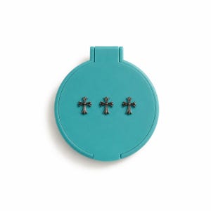 May include: A round, teal-colored compact mirror featuring a hinged top lid. The exterior surface is decorated with three small, metallic, silver-toned cross emblems arranged in a horizontal row across the center of the circular casing.