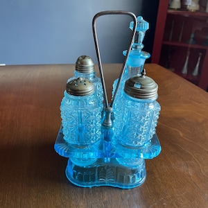 May include: A vintage blue glass condiment set with a metal handle. The set includes two salt shakers, a pepper shaker, and a cruet, all with ornate, textured glass. The set sits on a matching tray.