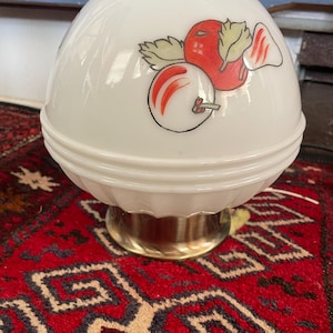 May include: A vintage white glass lamp with a brass base. The lamp shade features a colorful illustration of a red apple with green leaves. The lamp is sitting on a red patterned rug.