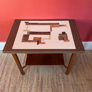 May include: A mid-century modern end table with a wood frame and a light pink top with a geometric design in brown and beige. The table has a lower shelf.
