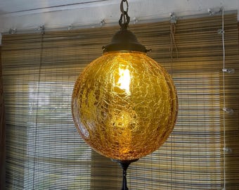 May include: A spherical pendant light fixture featuring a textured amber crackle glass globe. The lamp is suspended by a metal chain and includes a dark metal cap at the top and a decorative finial at the bottom, emitting a warm, glowing light.