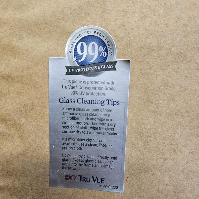 May include: A white label with a blue circle containing the text "Helps Protect From Fading 99%". The label also includes the text "UV Protective Glass", "This piece is protected with Tru Vue Conservation Grade 99% UV protection.", "Glass Cleaning Tips", and instructions for cleaning the glass. The label also includes the text "Tru Vue" and "M99-01230".