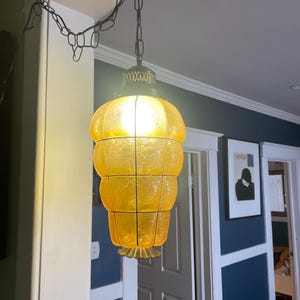 May include: A vintage, hanging pendant light with a unique, layered design. The light fixture features a warm, amber-colored glass shade with a crackle pattern, suspended by a black chain. The fixture is illuminated, casting a warm glow.