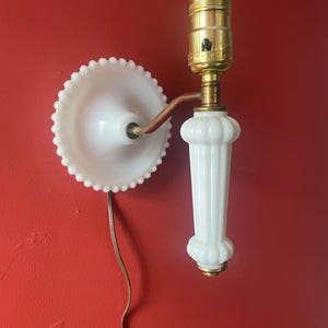 May include: A vintage white wall sconce with a brass fixture and a decorative, fluted base. The sconce features a white, scalloped shade and a brown electrical cord. The fixture is mounted on a red wall.