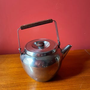 May include: A stainless steel teapot with a wooden handle and lid knob. The teapot has a rounded body and a spout for pouring. The handle is attached to the pot with metal brackets. The teapot is sitting on a wooden surface.