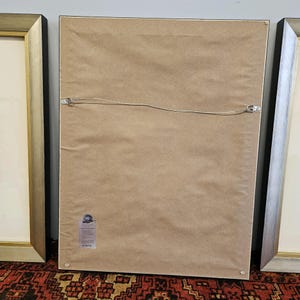 May include: The back of a framed picture with a brown paper backing. The frame is black with a gold trim. There is a small sticker on the bottom right corner of the backing.