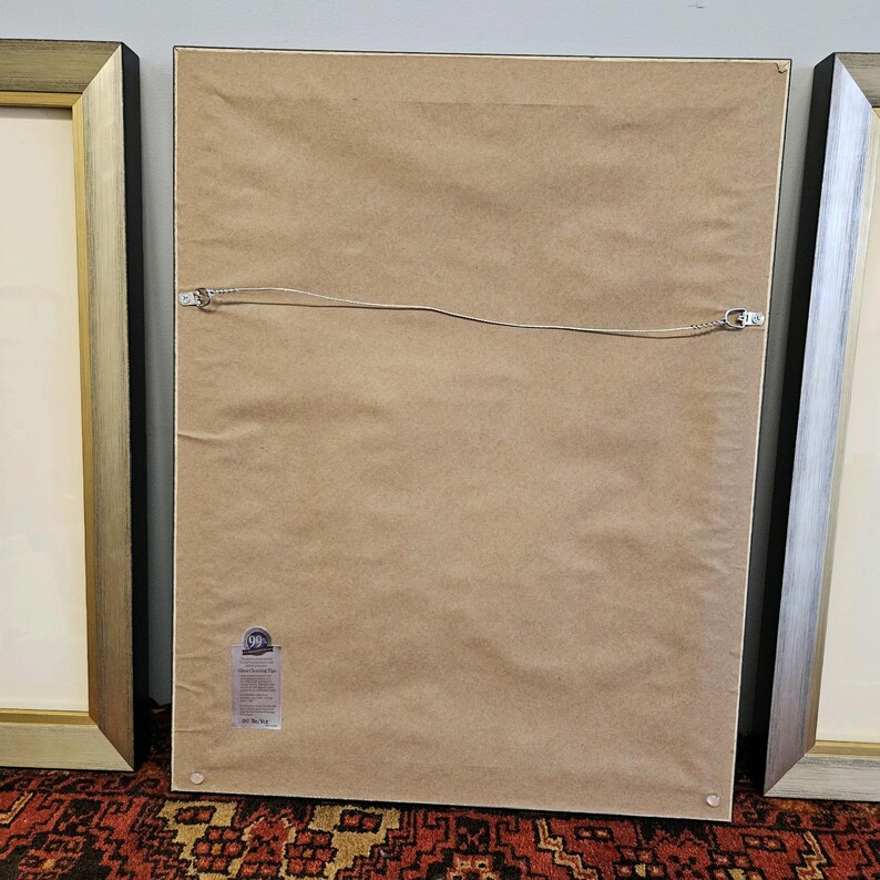 May include: The back of a framed picture with a brown paper backing. The frame is black with a gold trim. There is a small sticker on the bottom right corner of the backing.