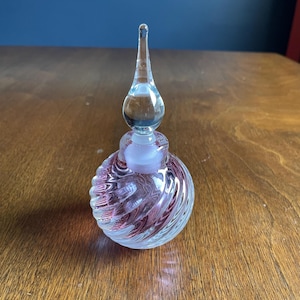 May include: A clear glass perfume bottle with a spiral design in shades of red and clear glass. The bottle has a long, teardrop-shaped stopper. The bottle is sitting on a wooden surface.