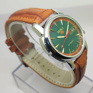 May include: A Seiko 5 automatic wristwatch featuring a round silver-tone stainless steel case, a textured green dial with gold-tone indices, and a day-date window. The watch includes a red second hand and a tan leather strap with a crocodile pattern.