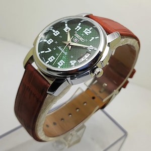 Könnte beinhalten: Eine Seiko 5 Automatikuhr mit einem runden Gehäuse aus silberfarbenem Edelstahl und einem strukturierten grünen Zifferblatt mit weißen arabischen Ziffern. Die Uhr verfügt über ein braunes Lederarmband mit Krokodilprägung und eine Datumsanzeige.