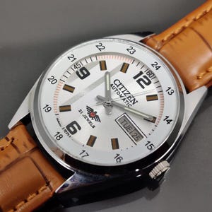 May include: A Citizen Automatic wristwatch with a silver dial, black Arabic numerals at 6 and 12, and a 24-hour outer track. The watch features a day-date window at the 3 o'clock position, silver hands, and a tan leather strap with a crocodile texture.