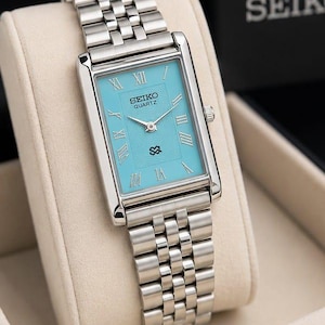 Könnte beinhalten: Eine rechteckige Seiko Quartz Armbanduhr mit einem helltürkisfarbenen Zifferblatt, silberfarbenen römischen Ziffern als Stundenmarkierungen und einem silberfarbenen Edelstahl-Gliederarmband. Das Zifferblatt zeigt das Seiko Quartz Logo.