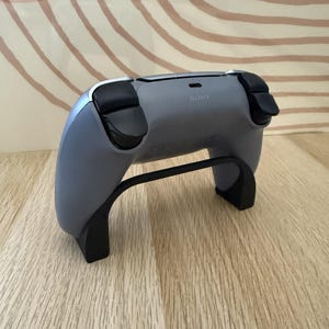May include: A grey wireless video game controller featuring black shoulder buttons and triggers, with the word SONY printed in small letters near the top center port. The controller is resting on a black plastic stand that supports its base.