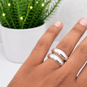 以下が含まれることがあります： 指に重ねて装着された、滑らかで光沢のあるシルバー調のリング2点。リングは丸みを帯びたドーム型のプロファイルで、光沢のある金属仕上げが施されています。バンドは装飾のないシンプルな形状で、全体を通して一定の厚みがあります。