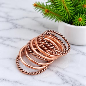 Peut inclure: Un ensemble de bagues empilées avec une finition en cuivre métallique. La collection comprend plusieurs anneaux fins, certains présentant une surface lisse et unie, d'autres conçus avec une texture torsadée en corde, disposés enchevêtrés.