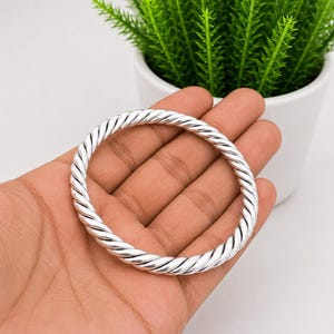 Puede incluir: Un brazalete rígido de tono plateado pulido que presenta un diseño continuo de cuerda trenzada. La banda metálica circular tiene una superficie lisa y reflectante con una textura diagonal repetitiva que crea un aspecto uniforme.