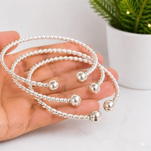 May include: Three silver-colored cuff bracelets featuring a twisted rope-like texture and polished spherical ball ends. The bracelets are open-ended, allowing for adjustment, and display a bright, metallic finish throughout the entire design.