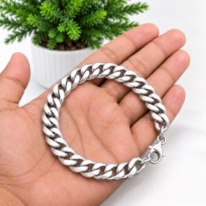 Puede incluir: Una pulsera de cadena tipo barbada en tono plateado pulido, con eslabones ovalados gruesos entrelazados y un cierre de mosquetón seguro. La pulsera presenta un acabado metálico brillante y un diseño uniforme en toda su longitud.