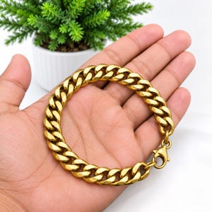 Puede incluir: Una pulsera de cadena tipo barbada en tono dorado con eslabones pulidos gruesos entrelazados y un cierre de mosquetón seguro. La pulsera está formada por una serie de eslabones metálicos redondeados y uniformes con un diseño de cadena clásico.