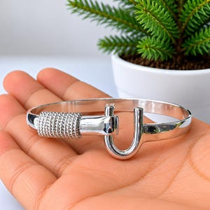 Peut inclure: Un bracelet jonc en métal argenté poli doté d'un fermoir en forme de U. Un côté du bracelet est orné d'un détail texturé en forme de corde enroulée qui entoure la bande, menant à une fermeture à crochet lisse et incurvée.