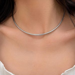 以下が含まれることがあります： 首の付け根にフィットする、滑らかで光沢のあるシルバー調の金属製チョーカーネックレス。ワイヤーのような丸みを帯びたデザインで、反射性のある金属仕上げが施された、継ぎ目のないクリーンな円形の形状が特徴です。