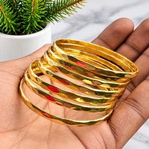 Könnte beinhalten: Ein Set aus fünf goldfarbenen Metallarmreifen mit einer gehämmerten, strukturierten Oberfläche. Jeder Armreif hat eine abgerundete, polierte Oberfläche, die das Licht reflektiert und eine Reihe sich wiederholender Vertiefungen erzeugt.