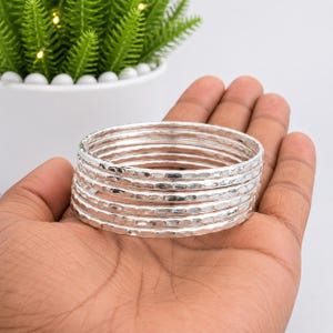 Puede incluir: Un conjunto de siete brazaletes apilables de plata, cada uno con una textura martillada distintiva que crea una superficie reflectante e irregular. Estas pulseras circulares son delgadas, metálicas y están diseñadas para usarse juntas.