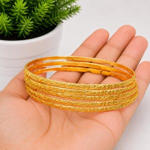 Peut inclure: Un ensemble de cinq bracelets joncs fins et circulaires en or, dotés d'un motif texturé répétitif sur la surface extérieure. Les bracelets sont empilés, présentant une finition jaune métallique brillante avec des détails gravés complexes.