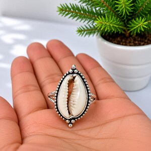 Peut inclure: Une bague en argent avec un coquillage cauri blanc central serti dans un cadre en métal perlé décoratif. La bague présente un design à anneau divisé et un sertissage ovale pointu qui souligne la texture naturelle et les stries du coquillage.