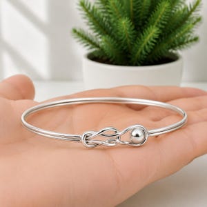 Puede incluir: Una pulsera rígida de tono plateado pulido que presenta una banda metálica fina y continua que forma un diseño de nudo estilo infinito en un lado, rematada con un pequeño abalorio esférico y liso al final de la estructura del nudo.